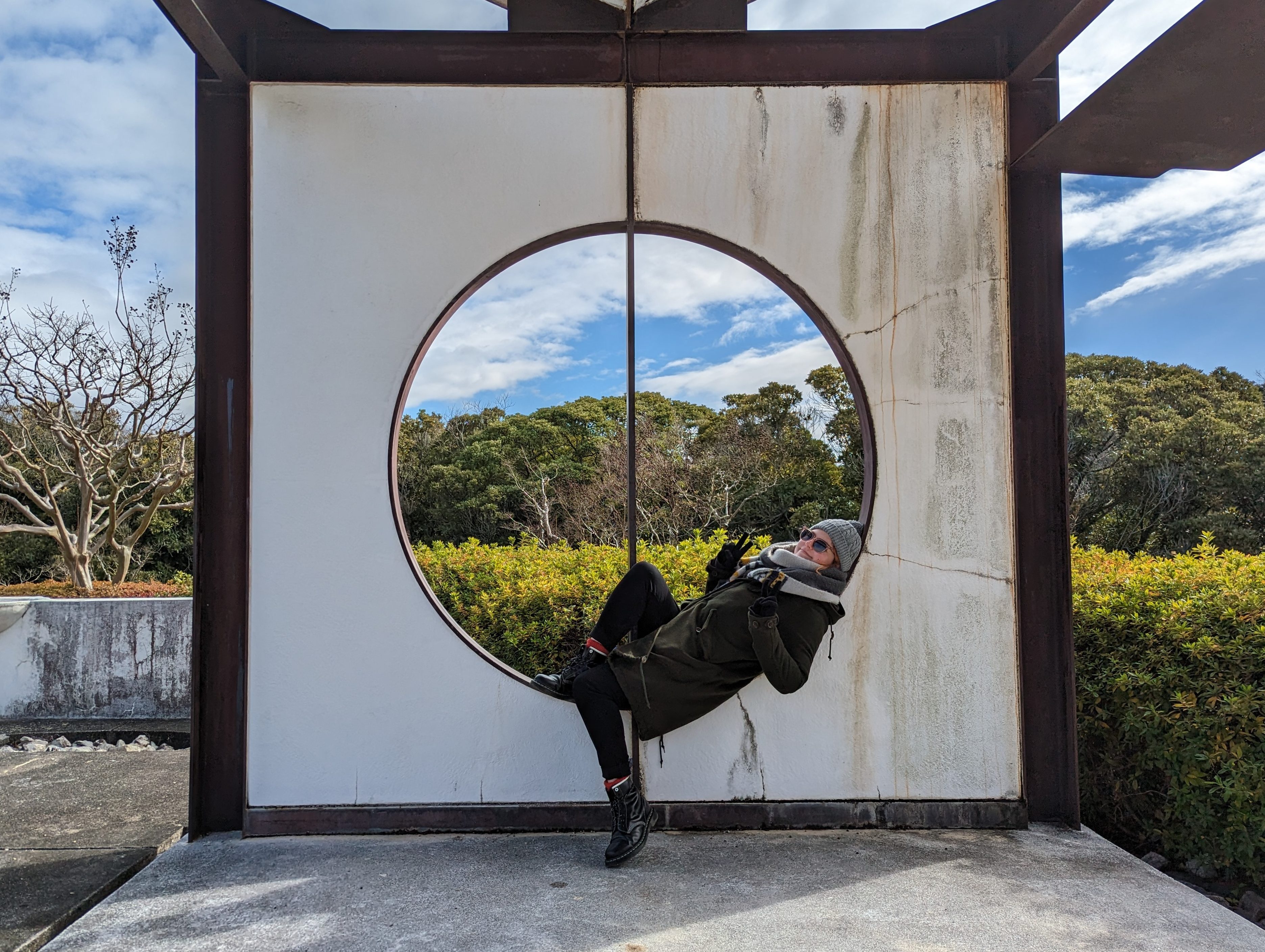 A woman smiling and posing in the large circular window-frame of an artistic structure in a park.